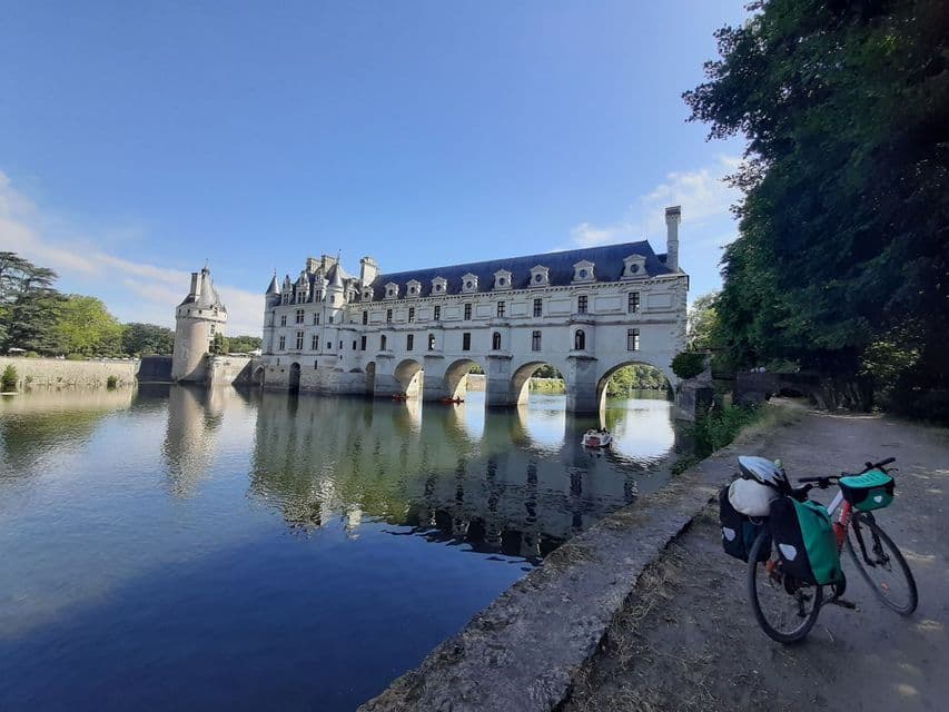 Deux vélos de randonnée garés sur un chemin de terre au bord d'une rivière, avec un grand château construit sur des arches au-dessus de l'eau en arrière-plan.