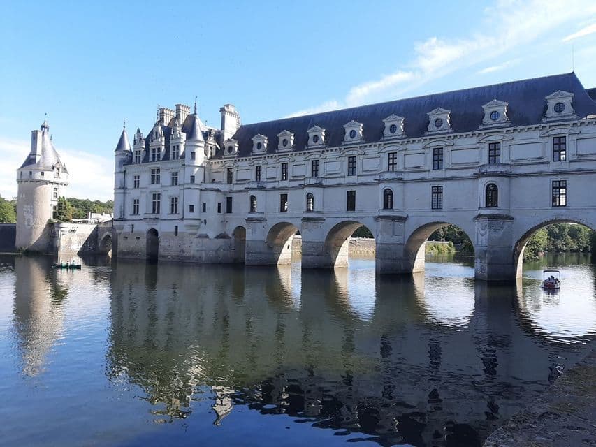 Un château en pierre blanche avec des arches enjambe une rivière calme, son reflet se mirant sur l'eau sous un ciel bleu clair.