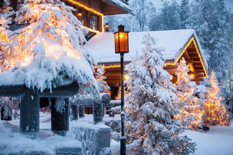 Una cabaña de madera y pinos cubiertos de nieve están decorados con luces de guirnalda brillantes junto a una farola encendida.