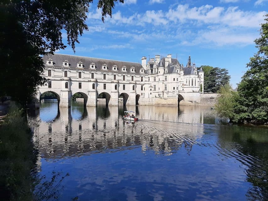 Un château blanc sur un pont arqué enjambe une rivière, avec son reflet dans l'eau et un petit bateau qui passe.