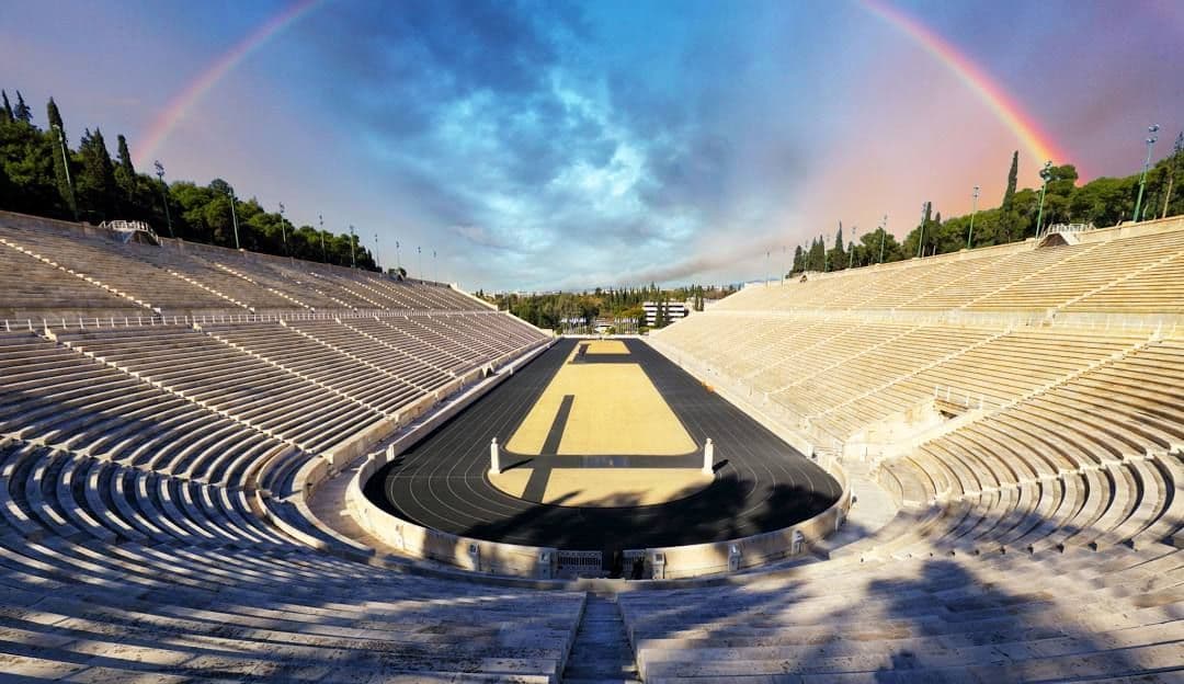 Una ripresa ampia di uno stadio in pietra vuoto con una pista da corsa, sotto un grande arcobaleno che si estende attraverso il cielo nuvoloso.