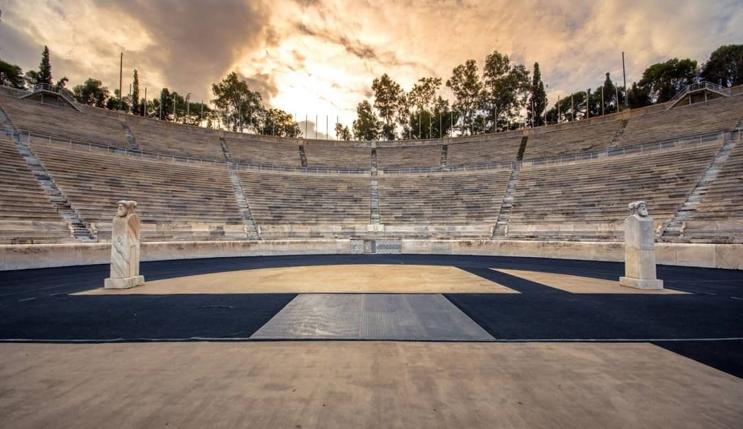 Uno stadio vuoto con gradinate in pietra, una pista di atletica e due statue sotto un cielo nuvoloso al tramonto.