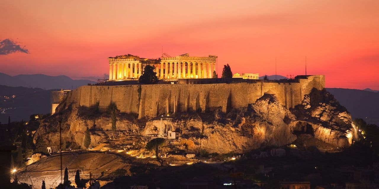 L'Acropoli illuminata si erge su una collina rocciosa contro un cielo infuocato di un tramonto rosso e arancione.