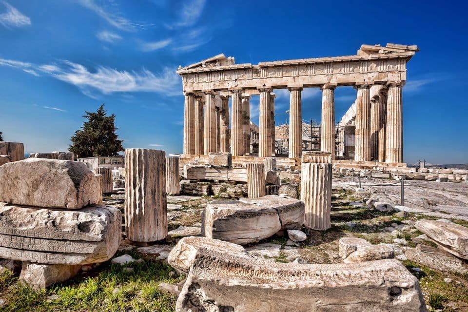 Un antico tempio di marmo con grandi colonne si erge su una collina, con rovine sparse in primo piano sotto un cielo azzurro.