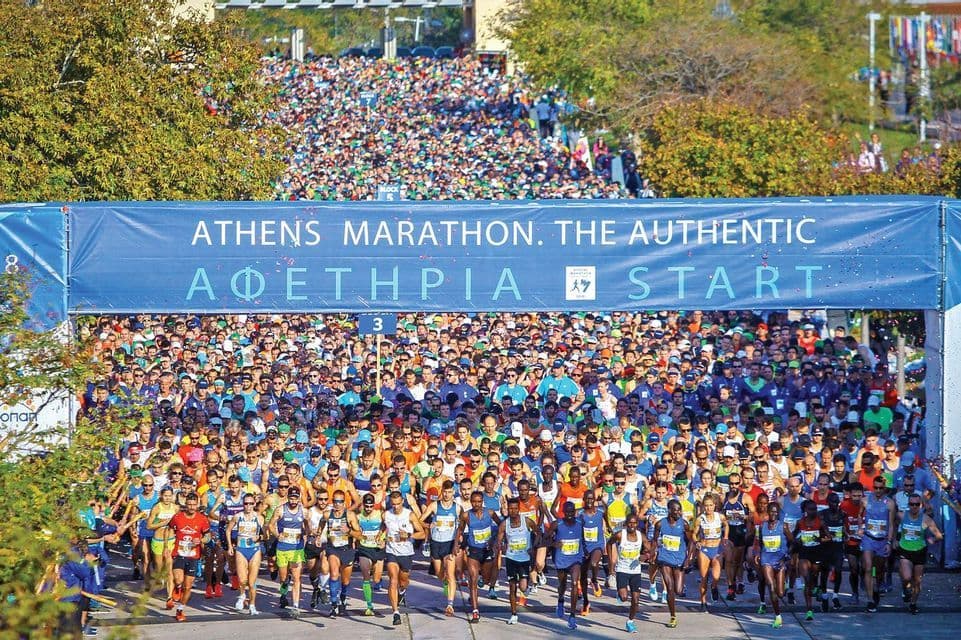 Un'enorme folla di corridori si riversa in avanti alla linea di partenza della Maratona di Atene sotto un grande striscione blu.