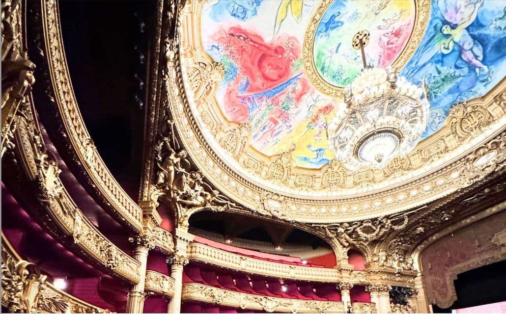Una vista dal basso di un teatro sontuoso, con una cupola del soffitto dipinta a colori vivaci e un grande lampadario di cristallo sopra balconi in velluto rosso.