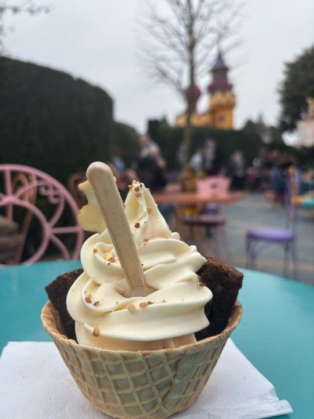 Nahaufnahme von Softeis in einer Waffelschale mit Brownie-Stücken und Nüssen, auf einem türkisfarbenen Tisch im Freien.