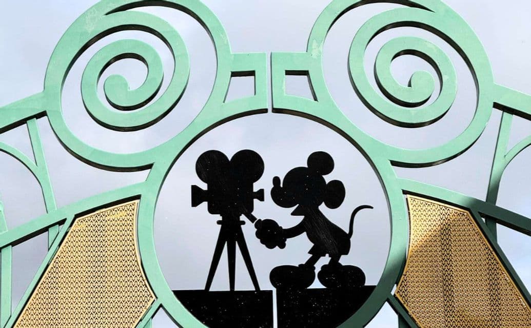 Primo piano di un cancello metallico verde con una sagoma nera di Topolino che manovra una cinepresa d'epoca.