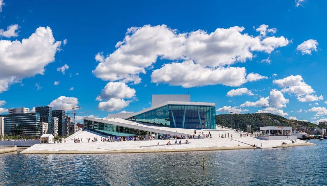 Ein modernes Gebäude mit einem schrägen weißen Dach und Glaswänden befindet sich an einer Uferpromenade unter einem strahlend blauen Himmel mit bauschigen Wolken.
