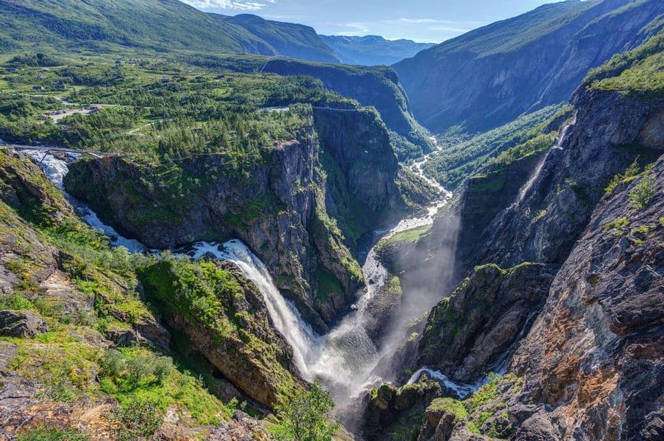 Una gran cascada cae en un cañón profundo, con un río que fluye por un valle montañoso exuberante y verde.