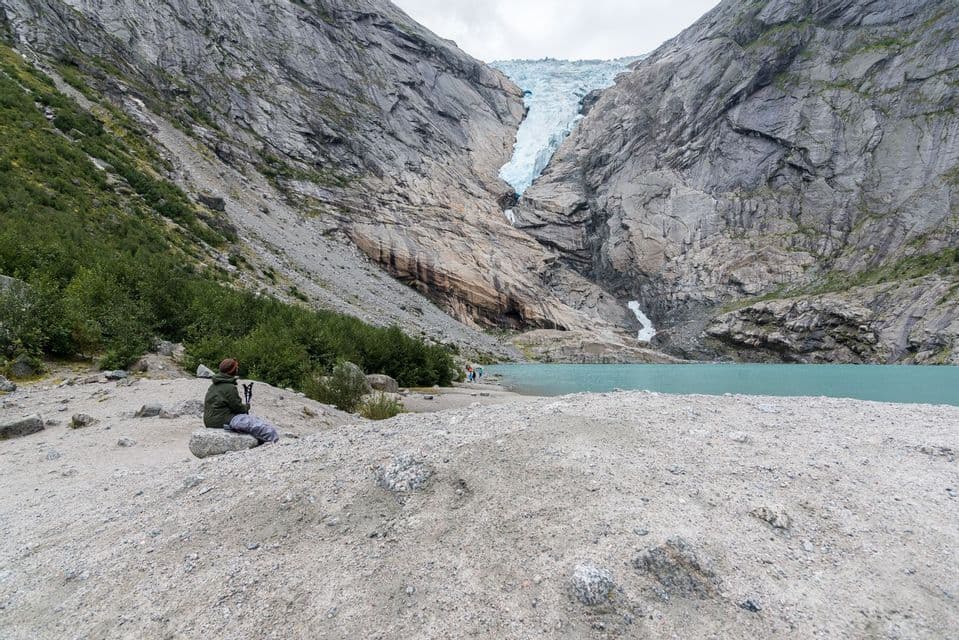 Un randonneur avec des bâtons de marche se repose sur un rocher dominant un lac glaciaire turquoise et un immense glacier de montagne.