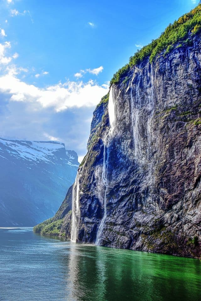 Des cascades dévalent une haute falaise verdoyante jusque dans un fjord, avec des montagnes enneigées sous un ciel bleu.