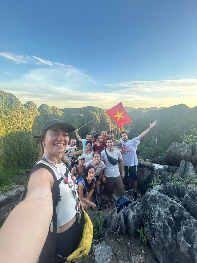 Eine WeRoad-Gruppe macht ein Selfie auf einem felsigen Berggipfel, hält eine vietnamesische Flagge und blickt auf grüne Karstberge.