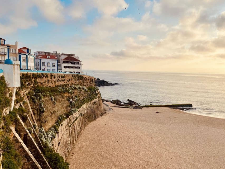 White buildings sit atop a fortified cliff overlooking a wide sandy beach and the ocean under a cloudy sky.