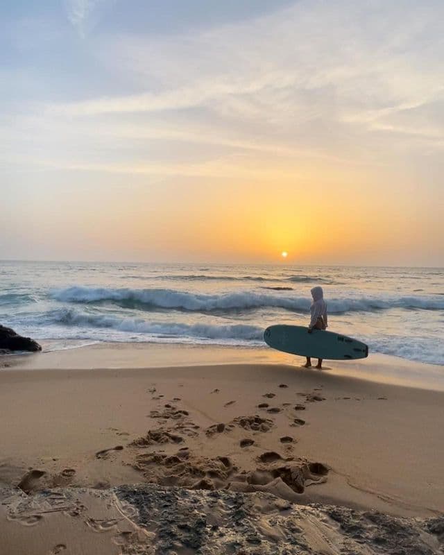 A person in a hoodie holds a surfboard while standing on a sandy beach, watching the waves at sunset.
