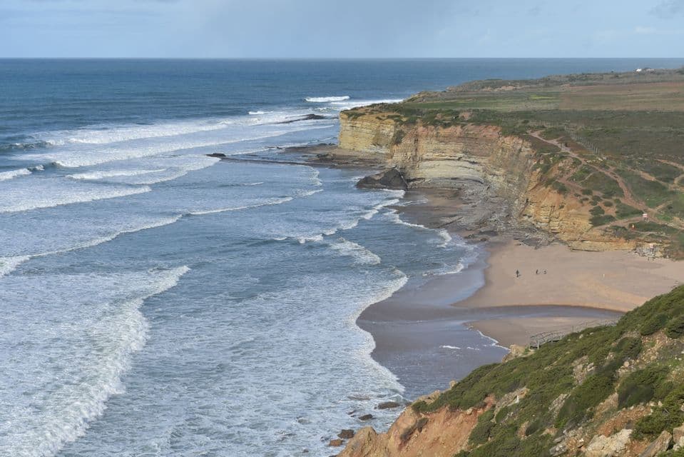 Veduta dall'alto di una costa con onde che si infrangono su una spiaggia sabbiosa accanto a scogliere alte e stratificate.