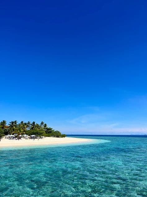Una vista dall'oceano cristallino e turchese verso una lingua di sabbia bianca con palme sotto un vasto cielo blu.