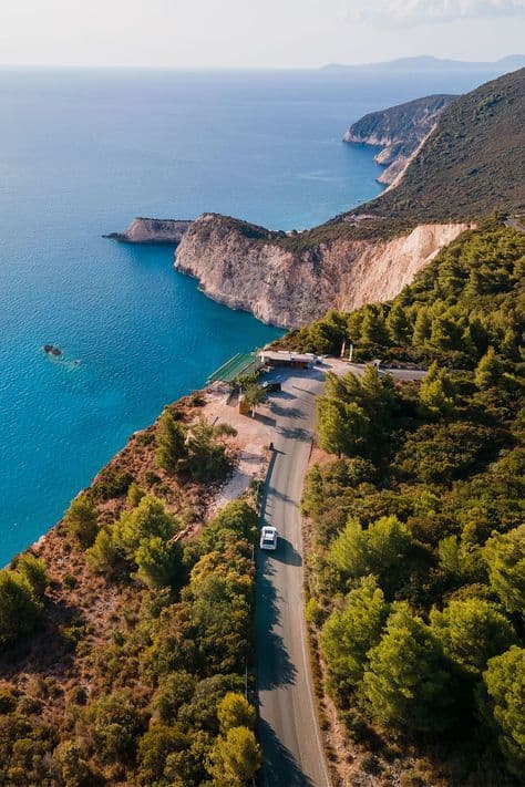 Veduta aerea di un'auto bianca che percorre una strada costiera tortuosa su una scogliera a picco sul mare blu.