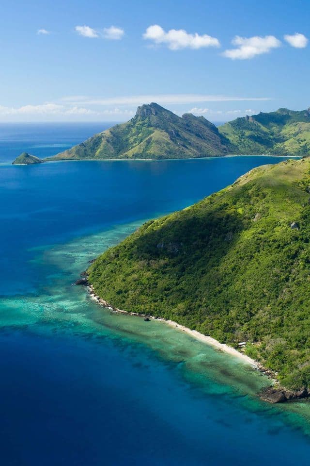 Una vista aerea di una lussureggiante costa di isola tropicale con acqua turchese e una barriera corallina sotto un cielo azzurro.