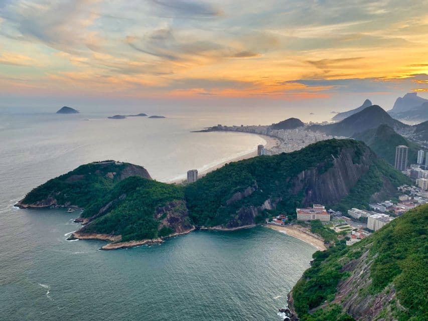 Vista panoramica di una città costiera con una lunga spiaggia curva e montagne rocciose verdi che incontrano l'oceano al tramonto.