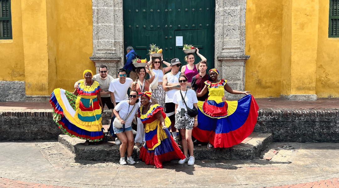 Un viaggio di gruppo WeRoad in posa su scalini di pietra con donne in abiti tradizionali colorati davanti a un edificio giallo.