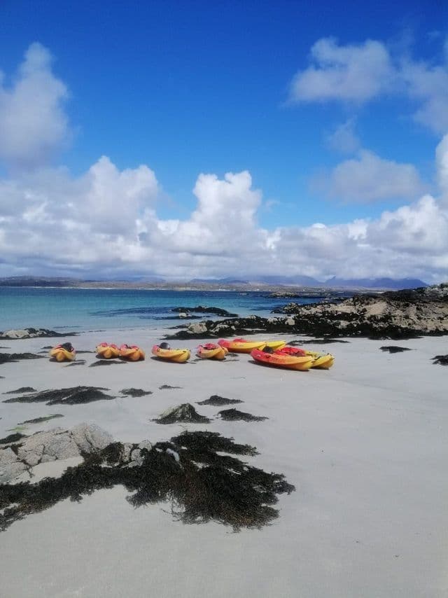 Varios kayaks amarillos y naranjas descansan en una playa de arena blanca con agua turquesa bajo un cielo parcialmente nublado.