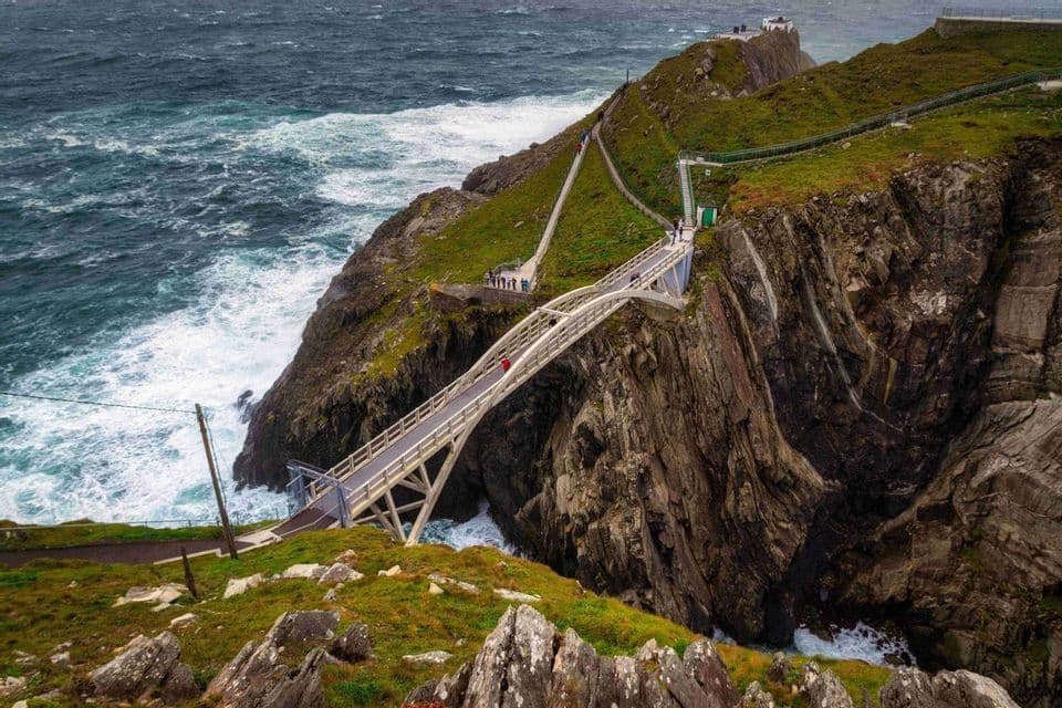 Un puente peatonal atraviesa un abismo entre dos acantilados empinados y cubiertos de hierba, mientras un mar turbulento rompe contra las rocas de abajo.