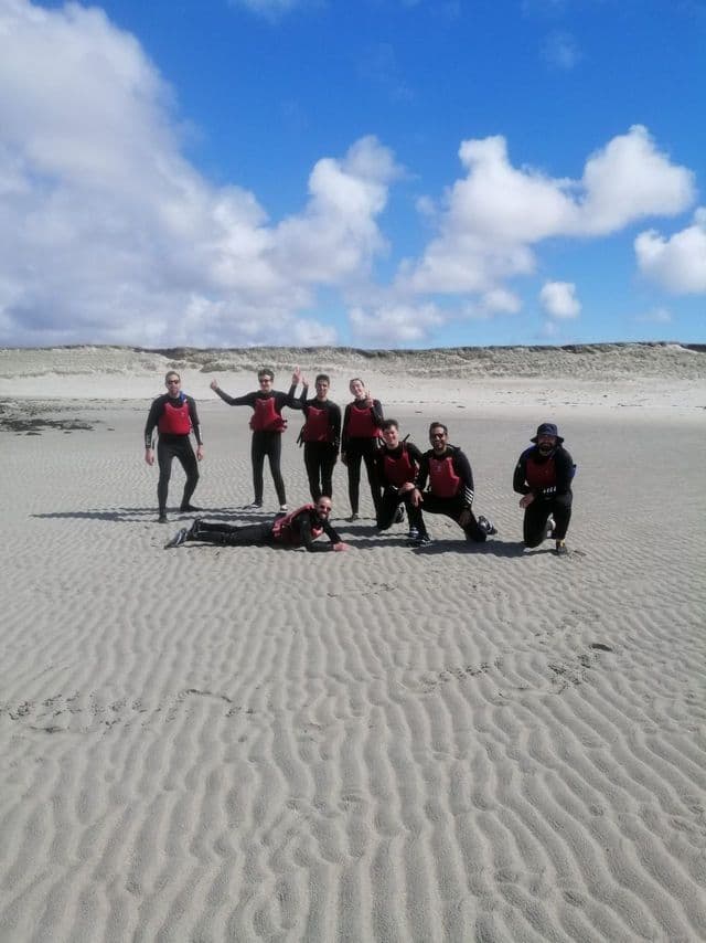 Un viaje en grupo de WeRoad con trajes de neopreno y chalecos salvavidas rojos posando para una foto en una playa de arena con dunas al fondo.