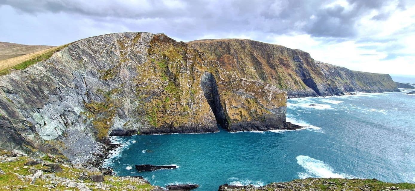 Acantilados escarpados y rocosos con un arco marino se precipitan en un océano turquesa bajo un cielo gris y nublado.