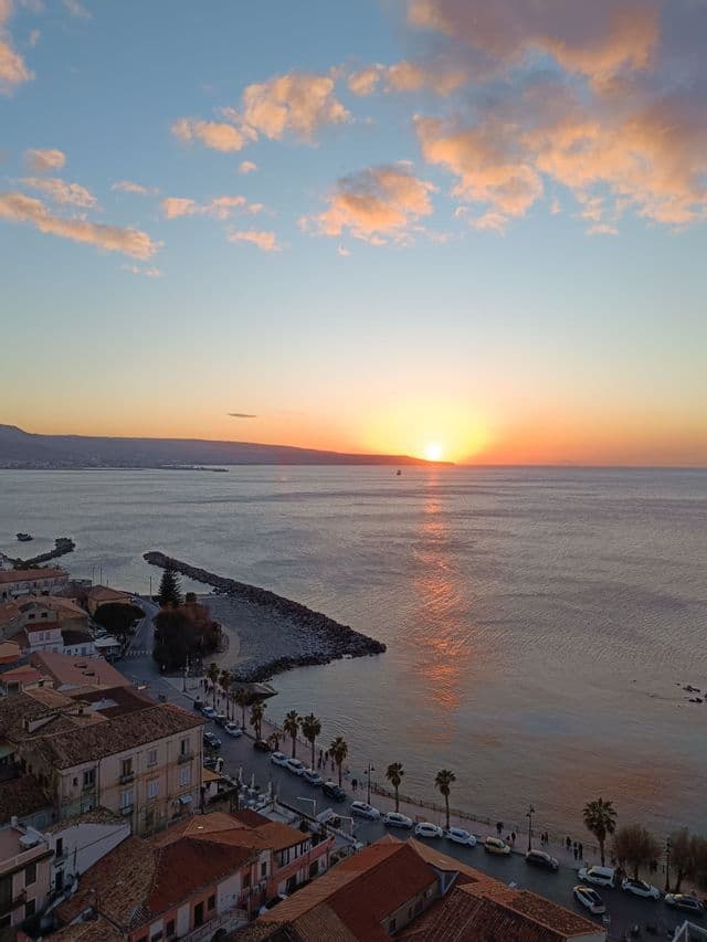 Vista aerea di una località costiera al tramonto, con un frangiflutti in pietra che si protende nel mare calmo.