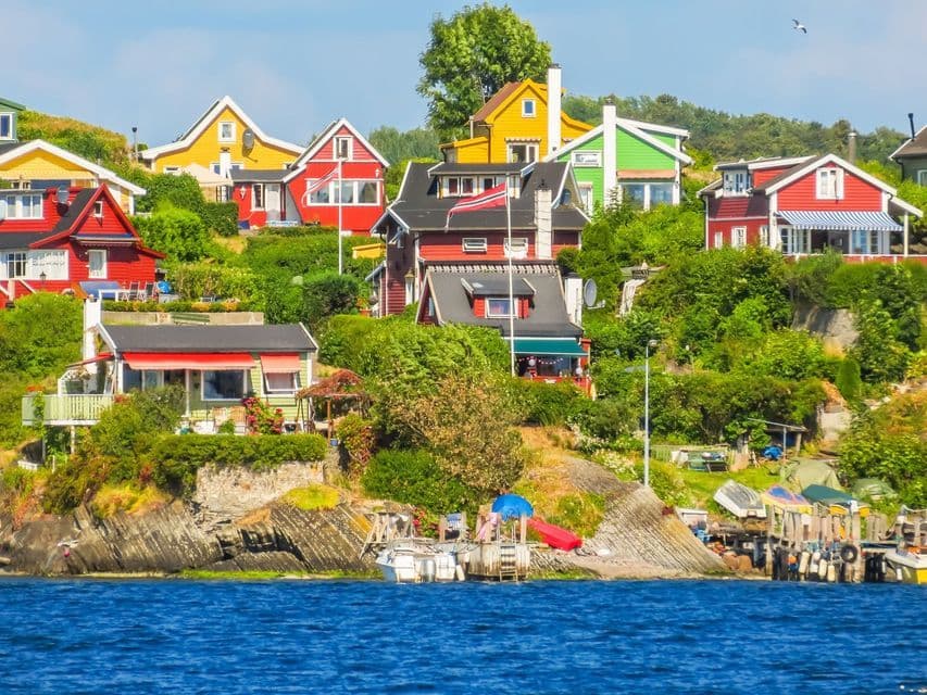 Des maisons en bois colorées sont construites sur une colline rocheuse verdoyante, au bord d'une étendue d'eau d'un bleu profond, avec des bateaux amarrés en contrebas.