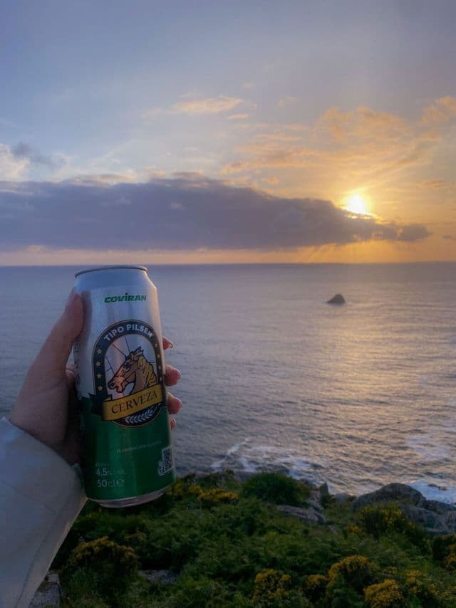 Una mano sostiene una lata de cerveza pilsner con vistas al océano desde un acantilado cubierto de hierba durante el atardecer.