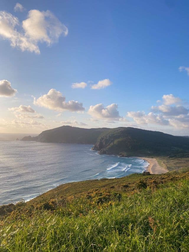 Una vista desde una colina cubierta de hierba con vistas a una bahía costera con una playa de arena, enmarcada por colinas verdes bajo un cielo parcialmente nublado.