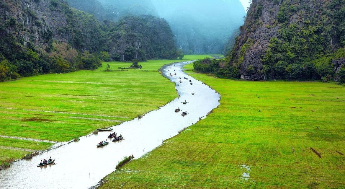 Eine WeRoad Gruppenreise in kleinen Booten navigiert einen gewundenen Fluss durch leuchtend grüne Felder, eingebettet zwischen großen, felsigen Bergen.