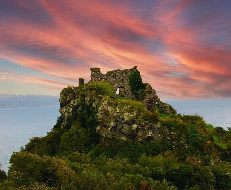 Rovine di un castello in pietra si ergono su una scogliera rocciosa e verde a picco sul mare, sotto un cielo con nuvole rosa e viola.