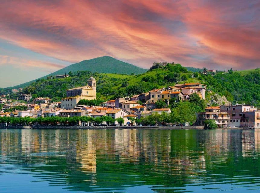 Un villaggio con una chiesa su un rigoglioso fianco di collina verde si riflette nell'acqua increspata di un lago sotto un cielo colorato al tramonto.