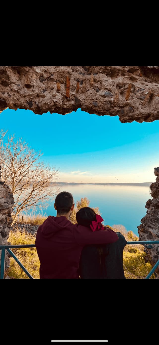 Una coppia, vista di spalle, si tiene a braccetto ammirando un grande lago da sotto un arco di pietra consumato.