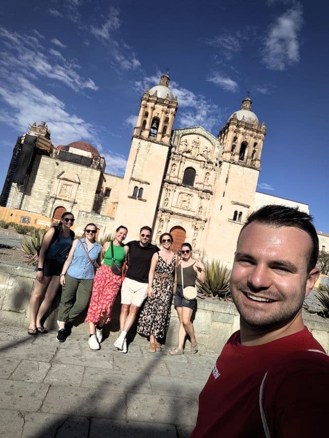 Ein Mann macht ein lächelndes Selfie mit seiner WeRoad-Gruppenreise vor einer alten Steinkirche unter blauem Himmel.