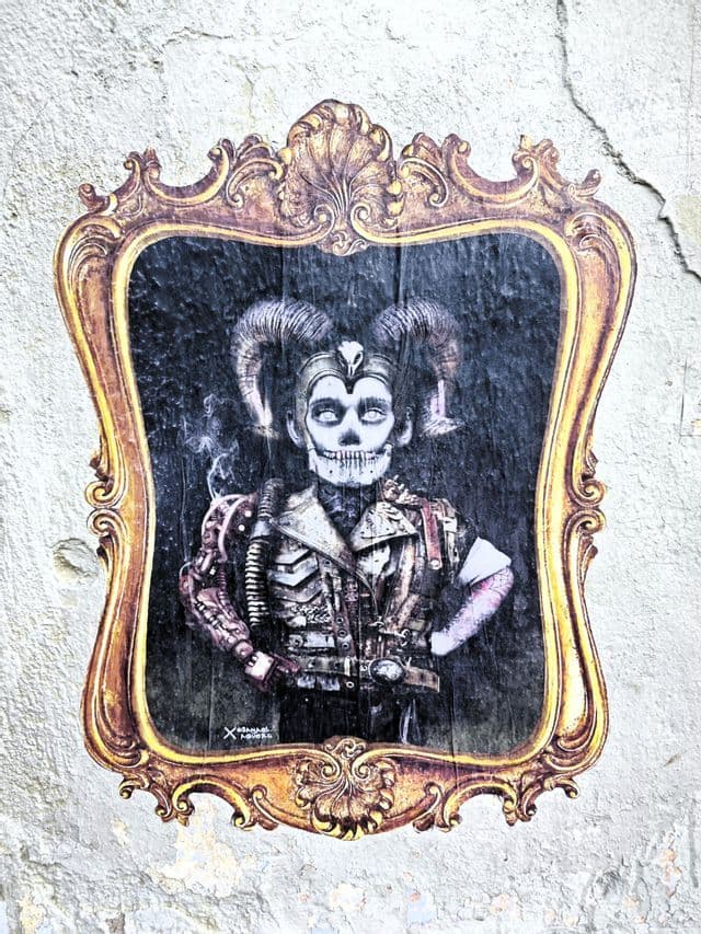 Streetart einer gehörnten, skelettartigen Steampunk-Figur in einem verzierten goldenen Rahmen, an eine rissige weiße Wand geklebt.