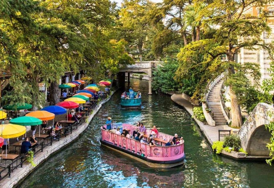 Two tour boats with passengers cruise down a canal next to riverside cafes with colorful umbrellas under lush green trees.