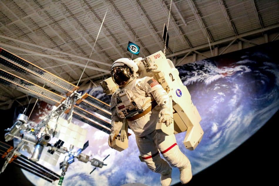 An astronaut spacesuit exhibit suspended in a museum, with a model of the ISS and an Earth backdrop.
