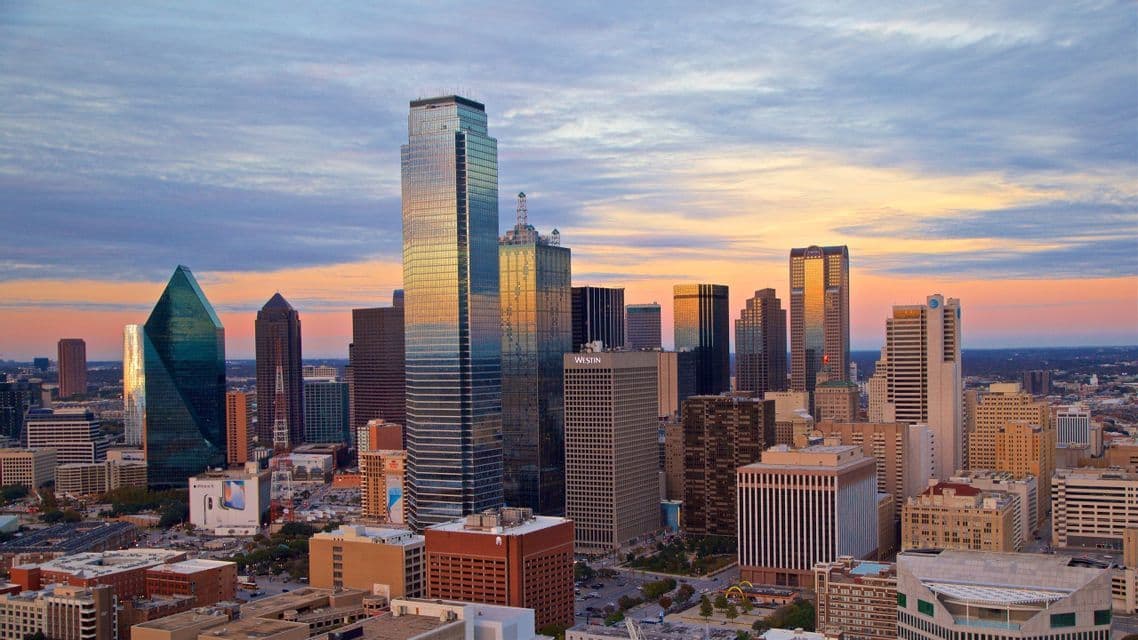 An aerial view of a dense city skyline with numerous skyscrapers reflecting the warm glow of sunset under a partly cloudy sky.