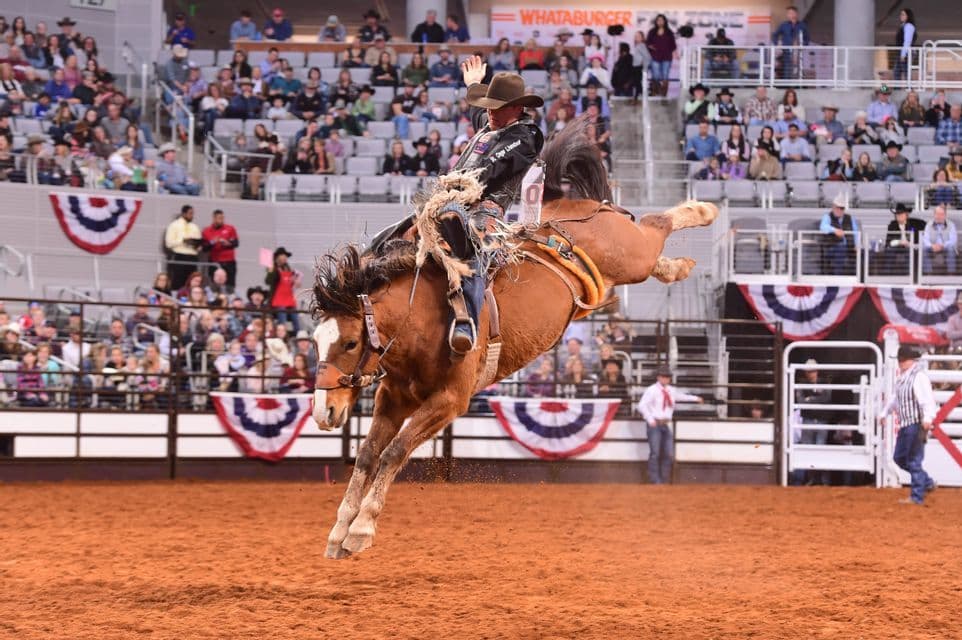 A cowboy in a hat rides a bucking horse mid-air in a rodeo arena, with one hand raised and spectators in the background.