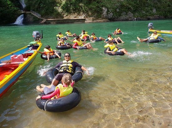 Un gruppo WeRoad in viaggio galleggia con giubbotti di salvataggio su ciambelle lungo un fiume cristallino, con una cascata sullo sfondo.