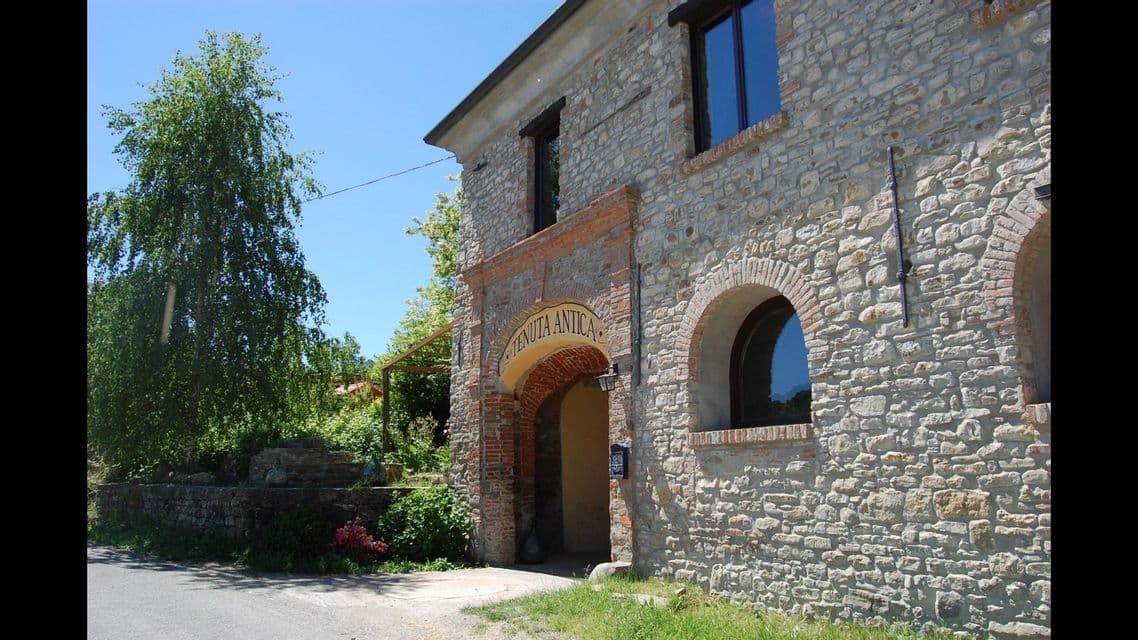 Un edificio rustico in pietra con un ingresso ad arco in mattoni etichettato 'Tenuta Antica', accanto a una strada alberata in una giornata di sole.
