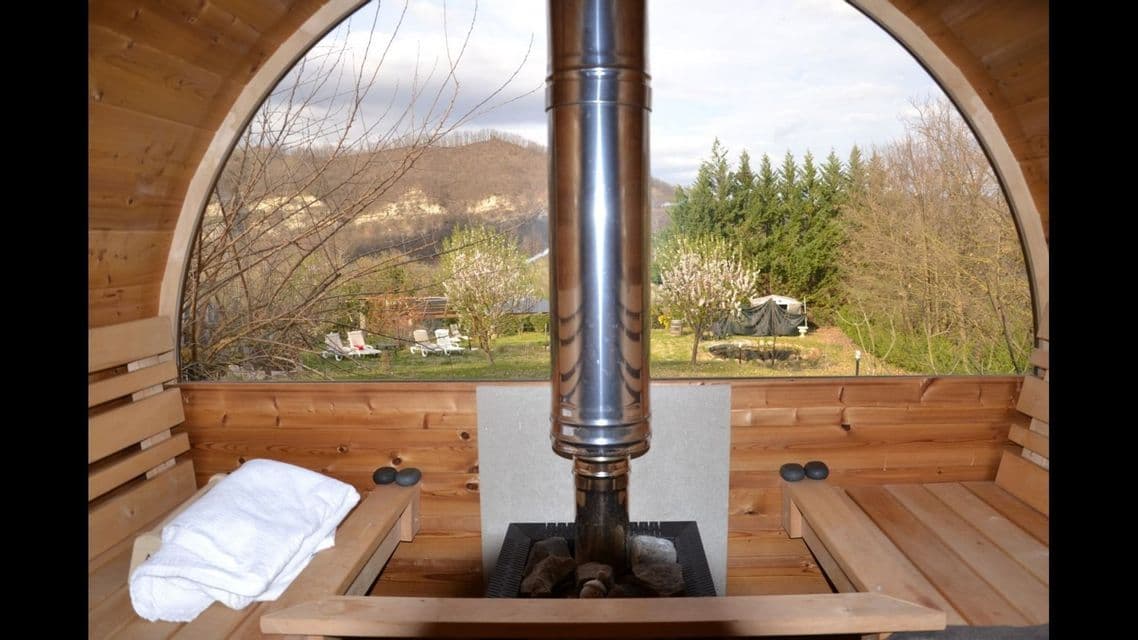 L'interno di una sauna a botte in legno con una stufa centrale, con una grande finestra che si affaccia su un paesaggio rurale con colline.