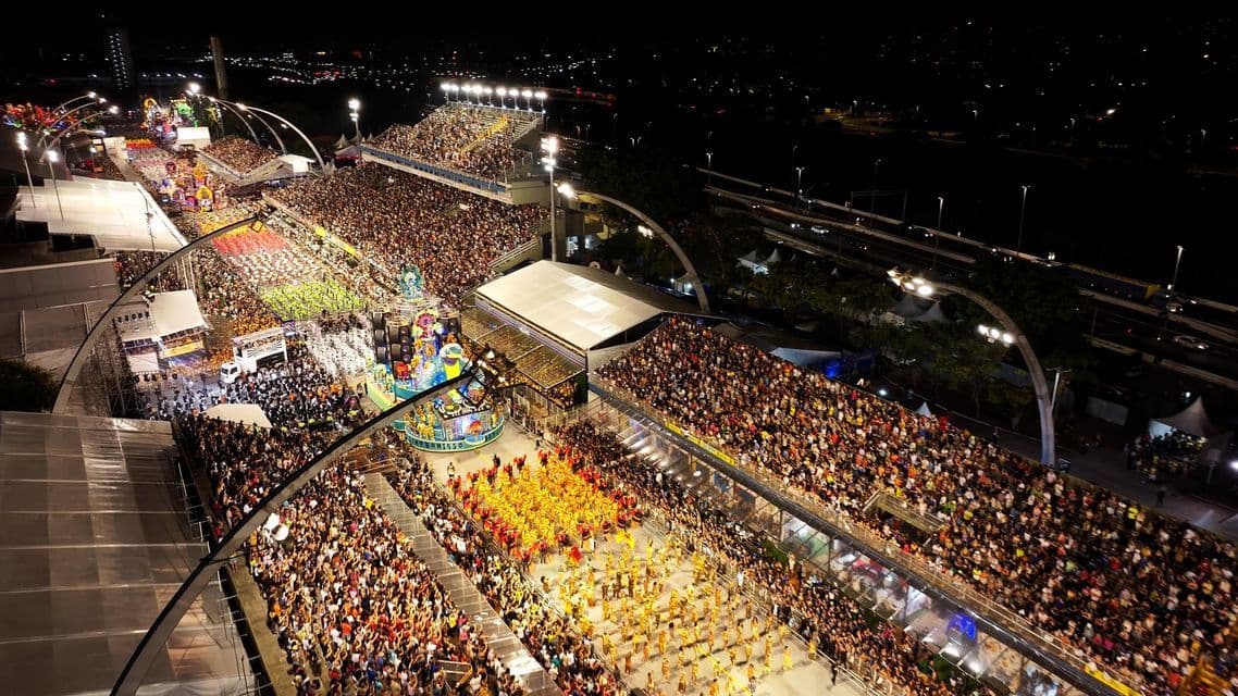 Una vista aérea de un vibrante desfile nocturno con carrozas coloridas y bailarines actuando para una gran multitud en las gradas.