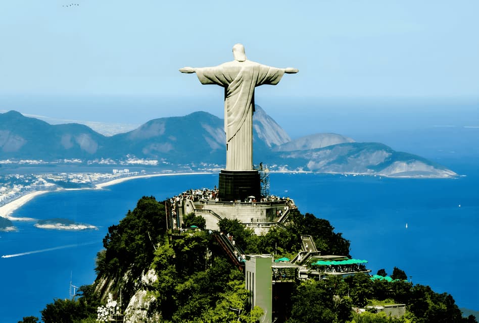 La statue du Christ Rédempteur, vue de dos sur une montagne, surplombant une baie avec un littoral urbain et des collines vertes au loin.