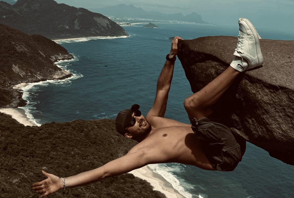 Un homme torse nu, portant casquette et lunettes de soleil, pose en suspension depuis un grand rocher, avec vue sur une côte pittoresque, une plage et l'océan.