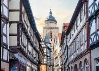 Una calle estrecha bordeada de edificios tradicionales con entramado de madera conduce a una gran torre de reloj de piedra a lo lejos.